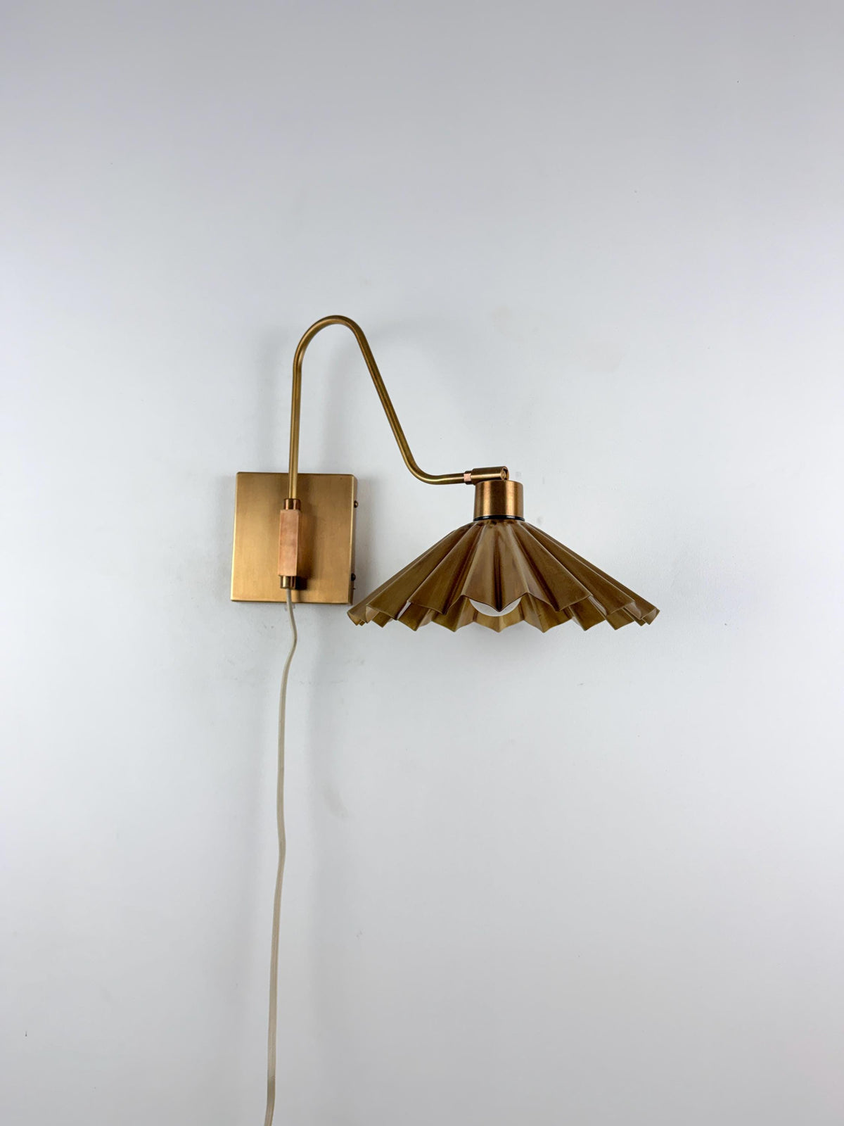 A mid-century modern wall sconce with a brass-finished wall plate, curved arm, and pleated shade, shown mounted on a plain white wall.