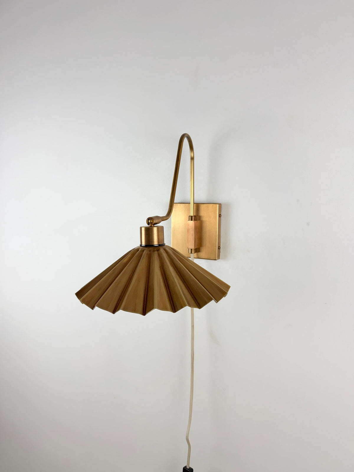 A mid-century modern wall sconce with a pleated metal shade and a curved arm, finished in a warm brass or bronze color, mounted on a plain white wall.