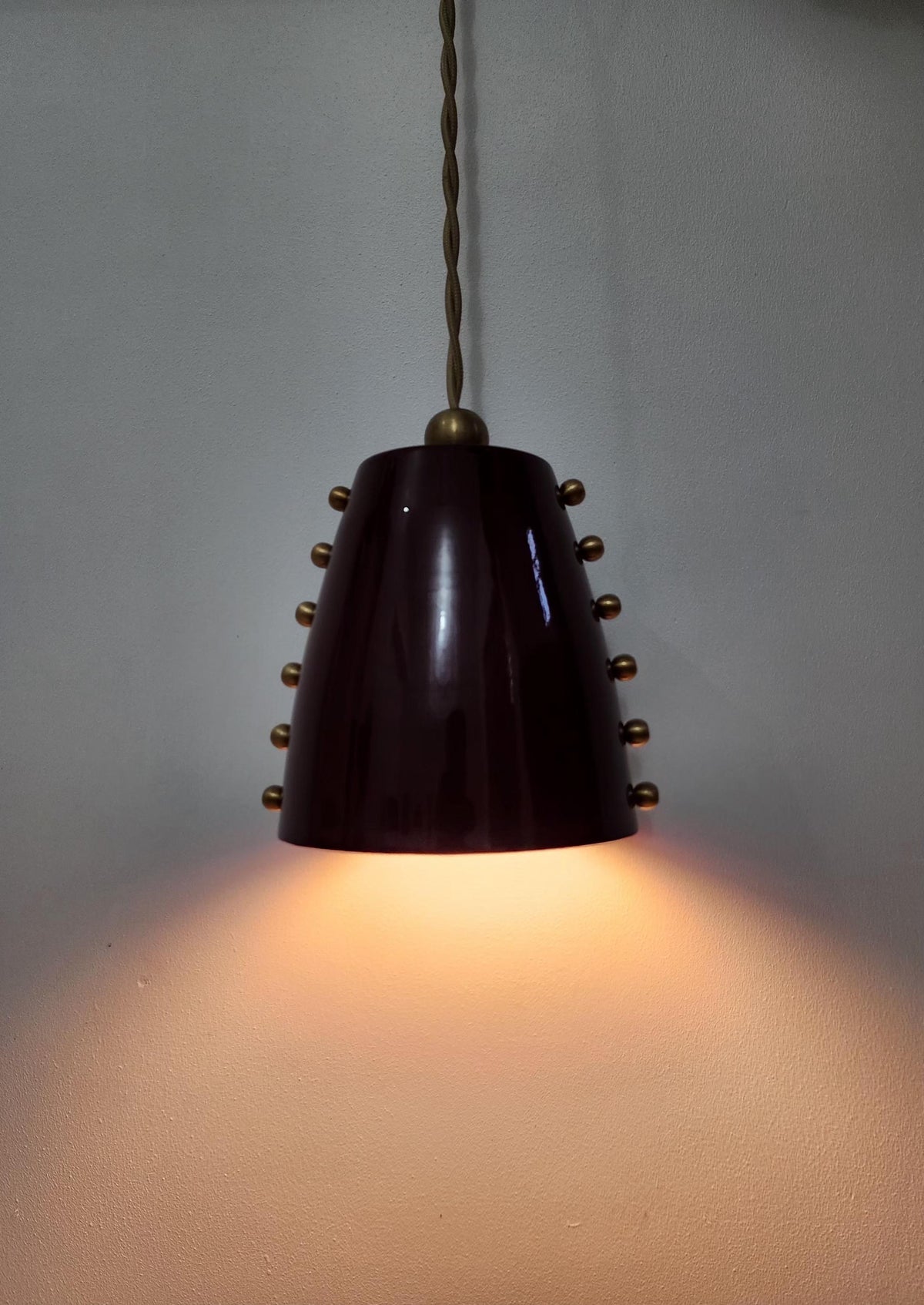 A vintage-style pendant light with a dark, glossy conical shade decorated with small metallic studs, suspended by a twisted metal cord against a plain wall with warm light glowing from below.