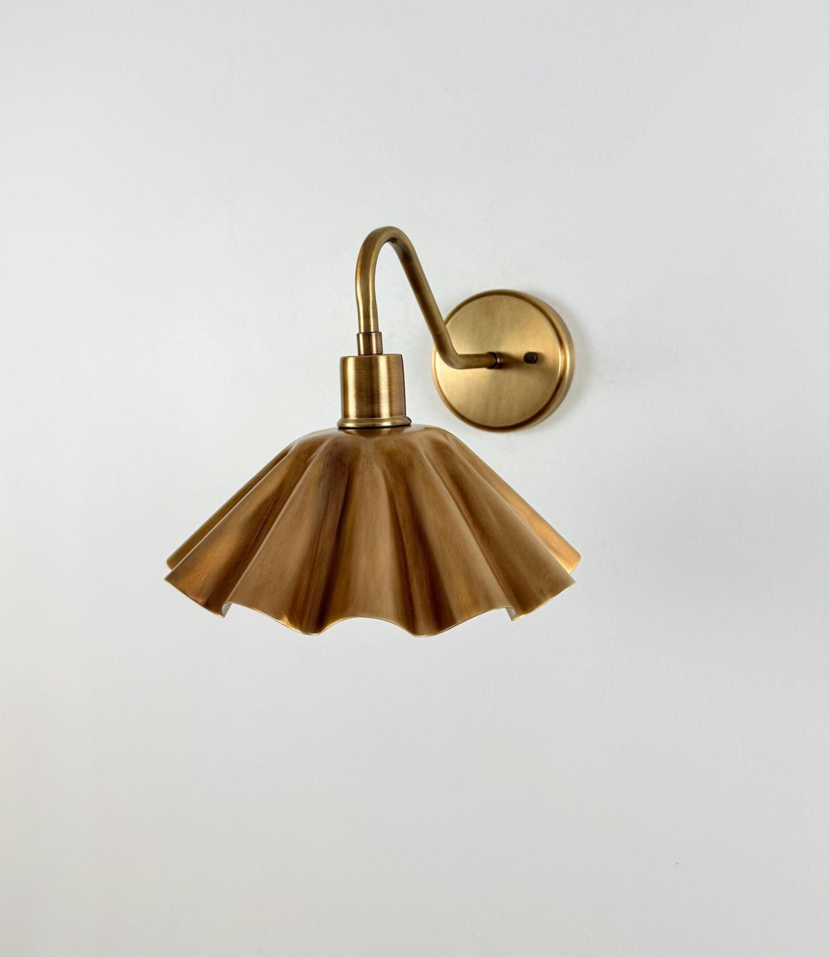 A vintage-style wall sconce with a fluted, pleated shade in a warm brass or bronze finish, mounted on a matching round wall plate with a curved arm, shown against a plain light gray wall.