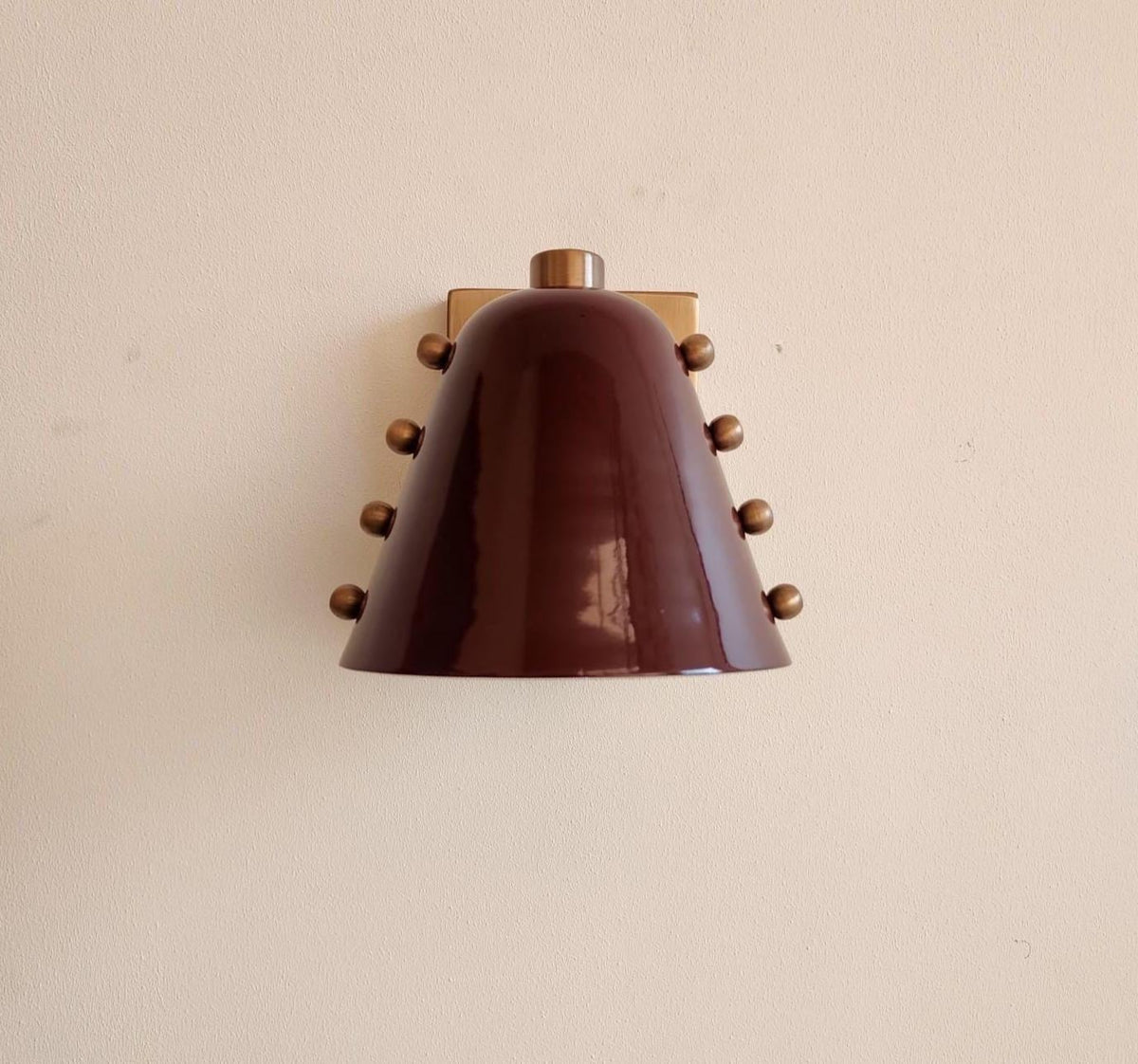 A vintage-style wall sconce with a glossy dark brown bell-shaped shade and small metallic bronze knobs, mounted on a plain beige wall.