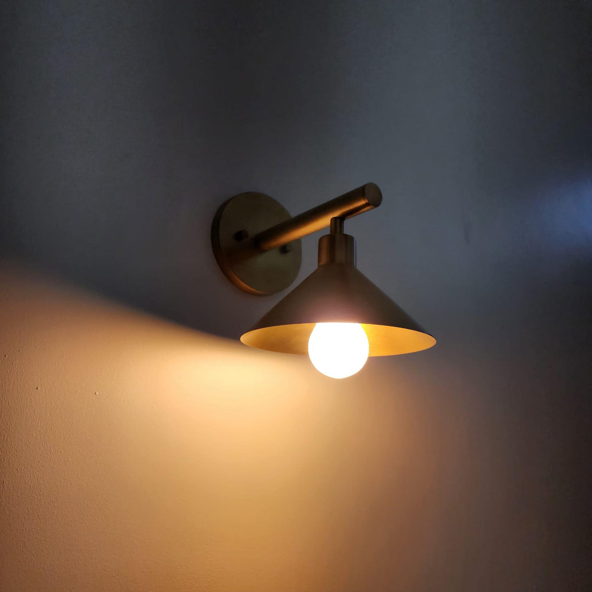 A wall-mounted industrial-style sconce with a warm brass finish, shown lit against a dark gray wall. The fixture features a circular backplate, a horizontal arm, and a conical shade housing a glowing light bulb.