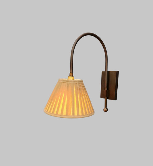 A wall-mounted sconce with a dark bronze curved arm, a rectangular wall plate, and a pleated conical shade glowing with warm yellow light, shown against a plain light gray background.